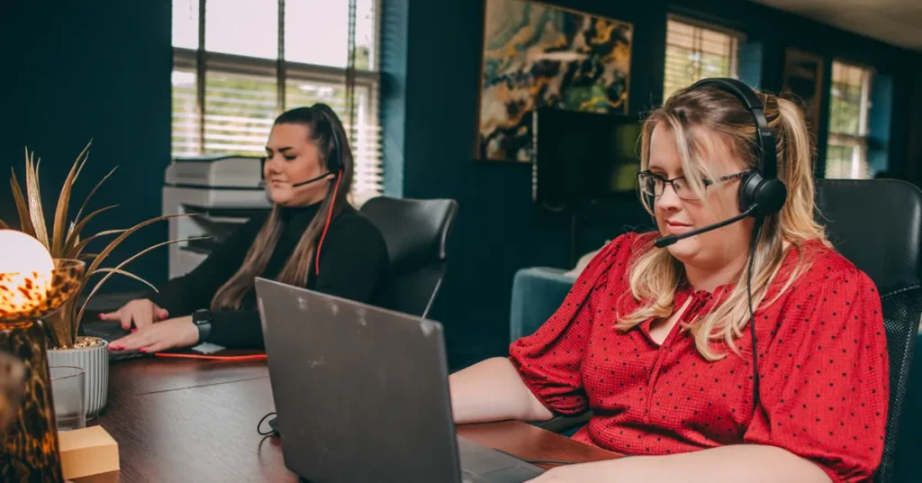 Two women sat next to each other at a desk with call answering headsets on